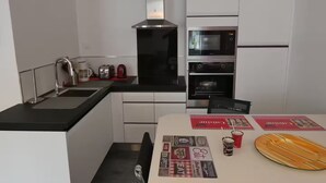 Een moderne keuken met witte kasten, een zwart aanrecht, een roestvrijstalen afzuigkap, een magnetron en een oven. Er liggen twee rode placemats op tafel.