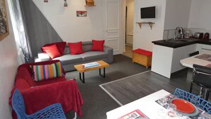 Een moderne woonruimte met een grijze bank, rode kussens, een houten salontafel, een blauwe stoel en een eethoek met een witte tafel en rode borden.