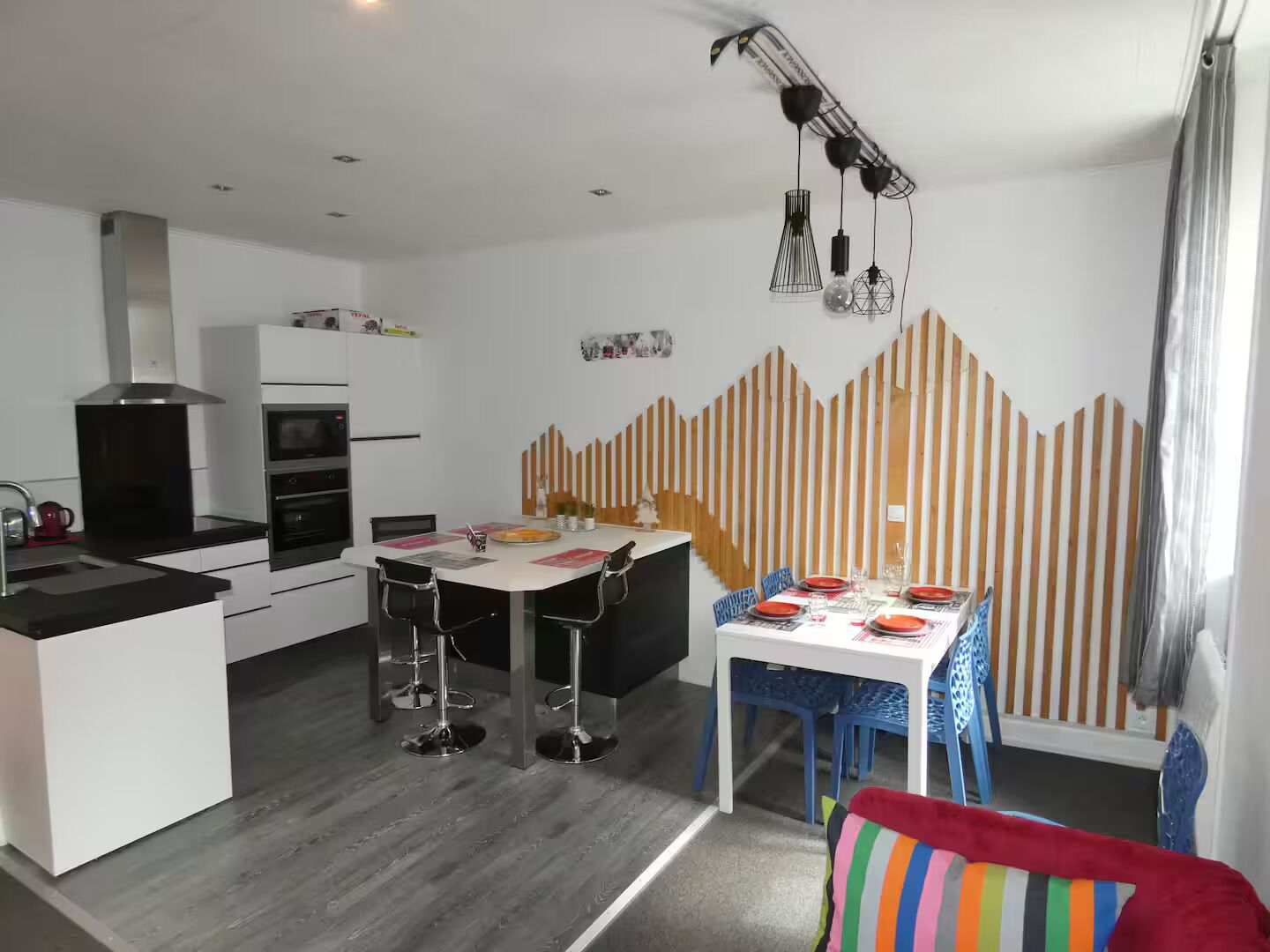 Eine moderne Küche mit weißen Schränken, ein Essbereich mit einem weißen Tisch und blauen Stühlen sowie ein farbig gestreiftes Kissen auf dem Boden.