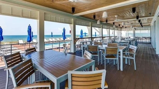 Ein überdachter Essbereich im Freien mit Holztischen und -stühlen mit Blick auf einen Strand.