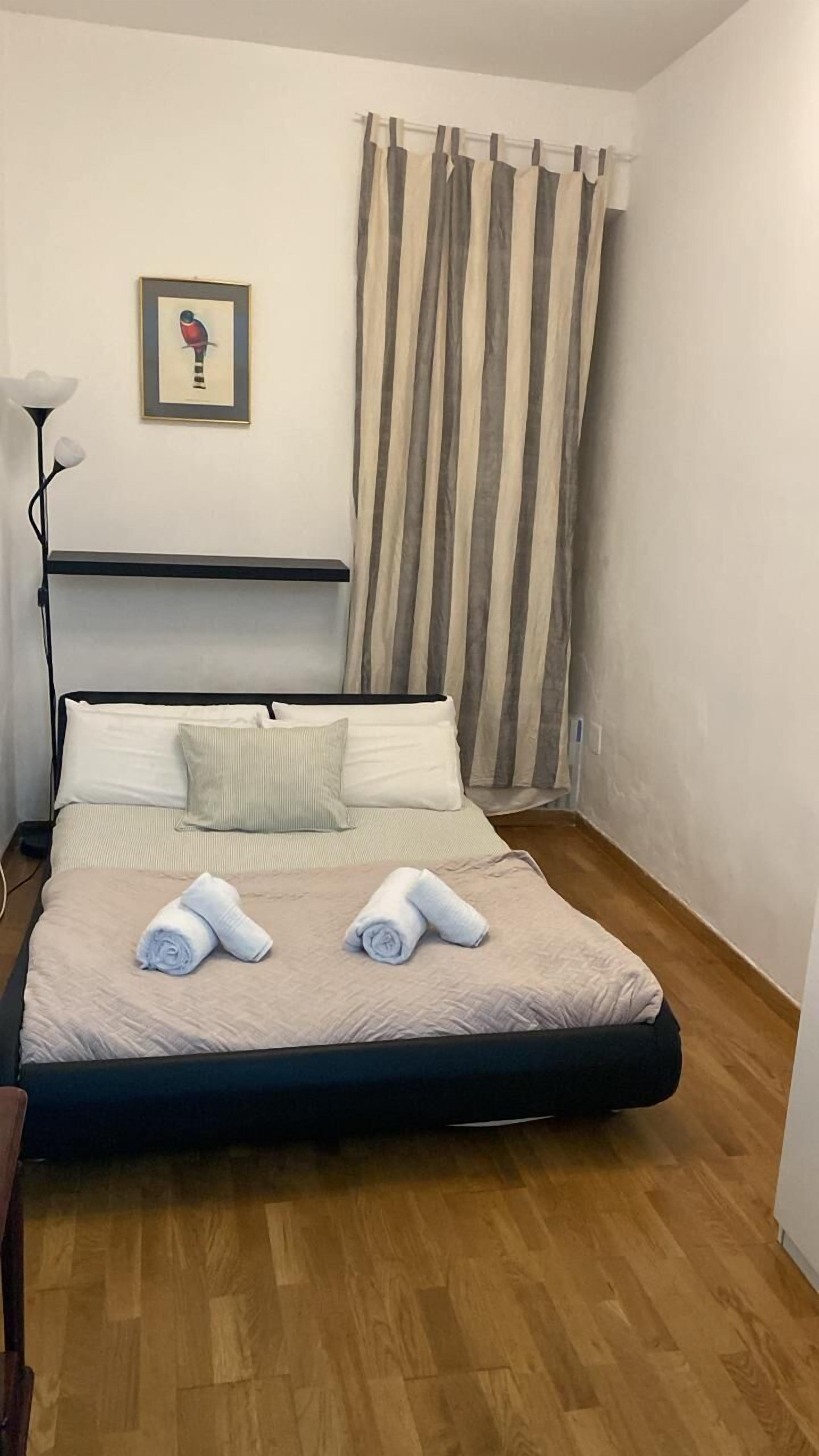 Una cama bien hecha con dos toallas enrolladas, un cuadro en la pared y una lámpara de pie.