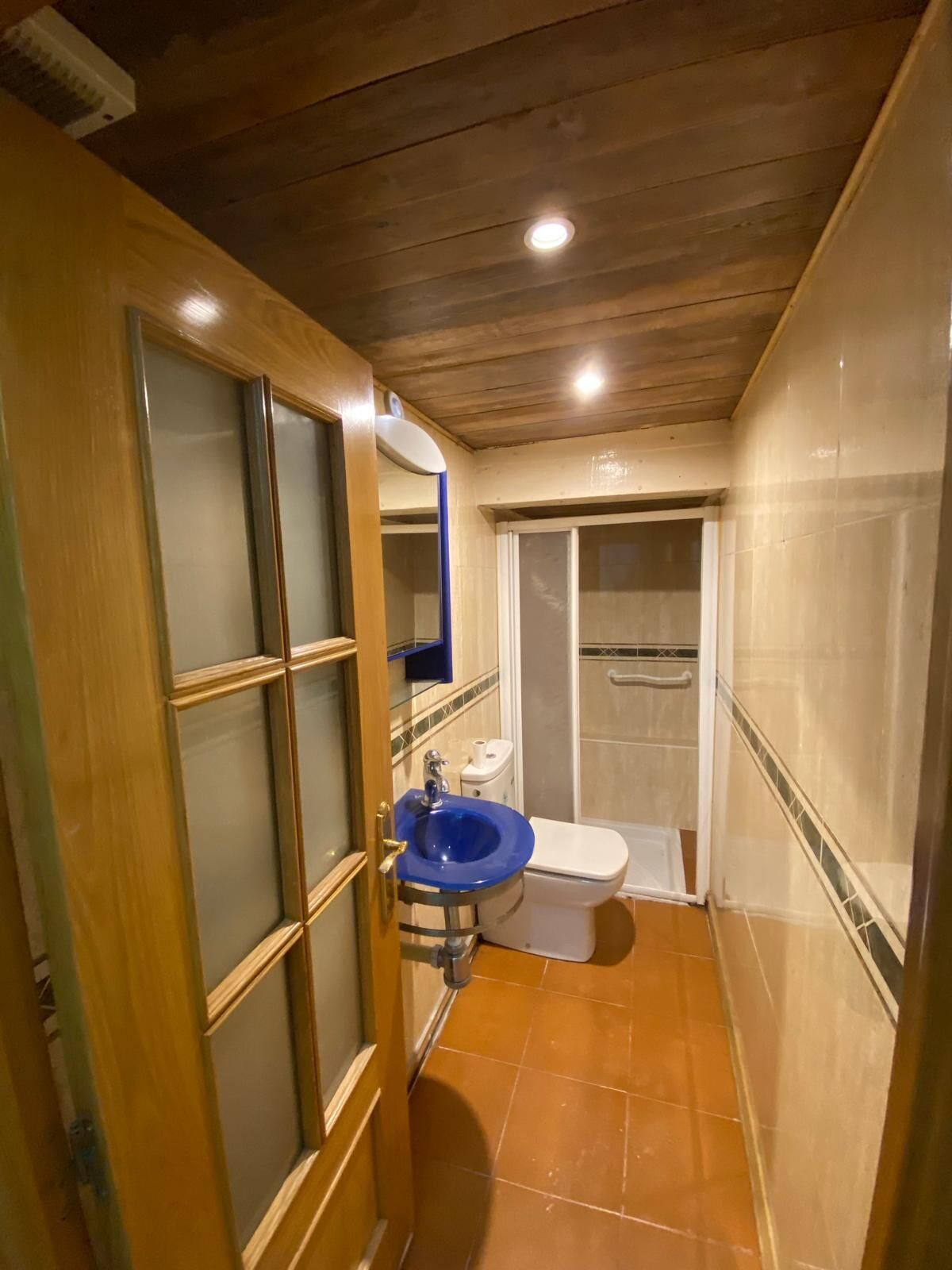 Un baño estrecho con puerta de madera, un lavabo azul, un inodoro y una zona de ducha.