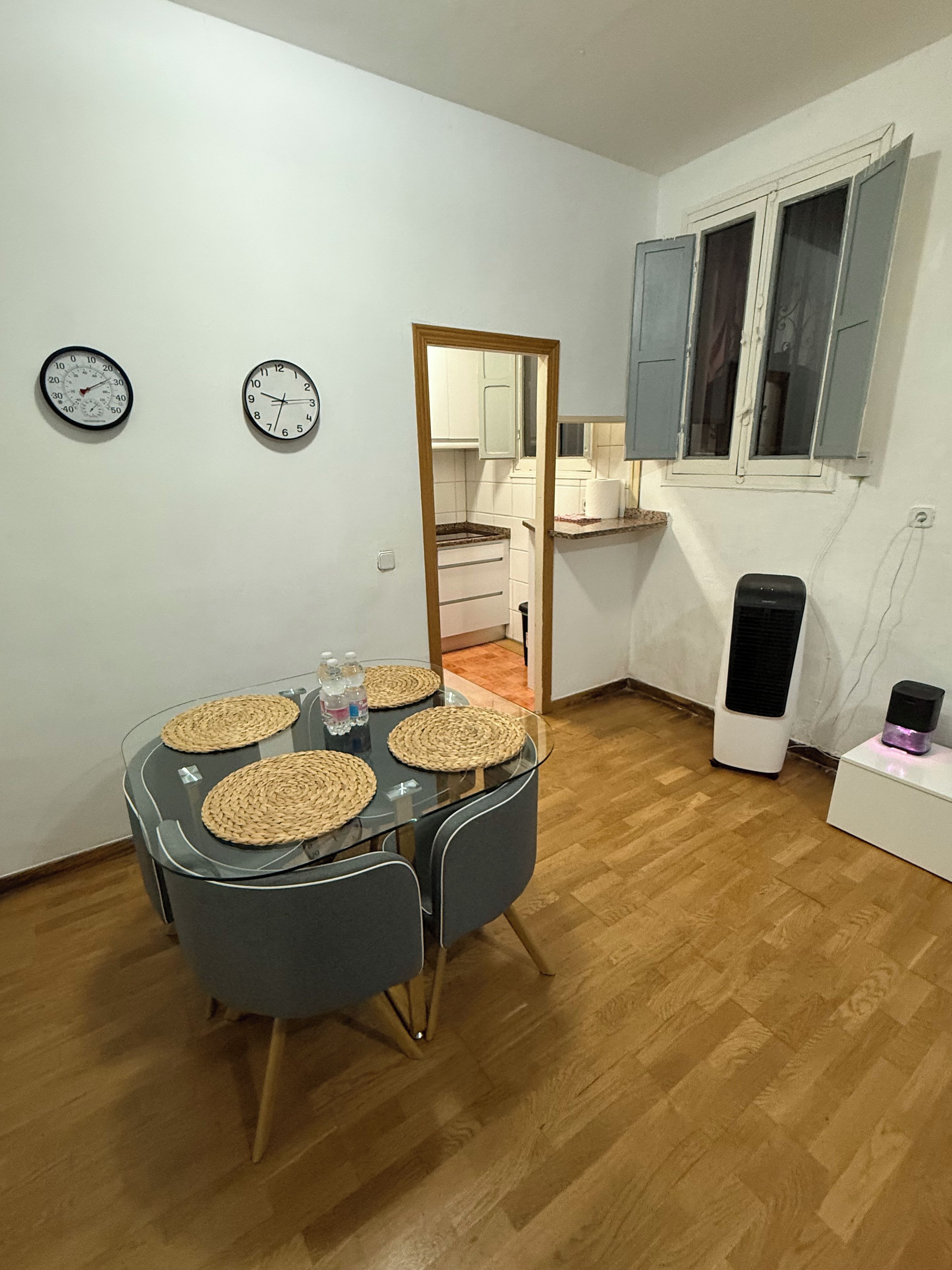 Cocina compacta con una mesa de comedor, dos relojes de pared, un espejo y una ventana con contraventanas.