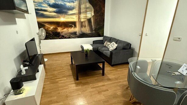 Un salon moderne avec un plancher en bois, un canapé gris, une table basse en verre et une grande fresque murale représentant un coucher de soleil sur une cascade.