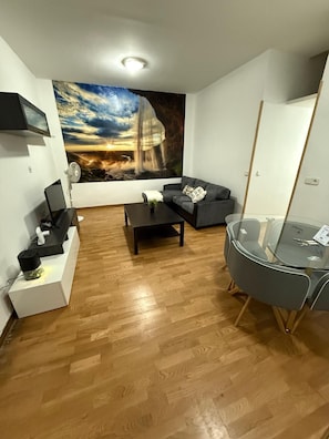 En moderne stue med trægulv, en grå sofa, et glas sofabord og et stort vægmaleri der viser en solnedgang over et vandfald.
