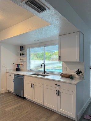 A modern kitchen with white cabinetry, a built-in dishwasher, a window with blinds, and a view of greenery outside. - Sunny Pool Getaway • Private Yard • Perfect Stay (Cape Coral)