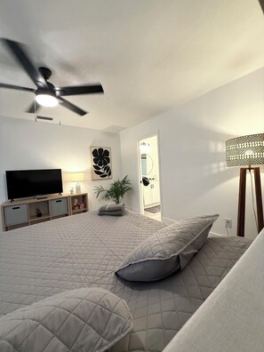A bedroom with a bed, a ceiling fan, a TV, a lamp, a plant, and a door. - Sunny Pool Getaway • Private Yard • Perfect Stay (Cape Coral)