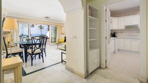 Uma sala de jantar com mesa e cadeiras, uma cozinha com armários brancos e vista para palmeiras pela janela.