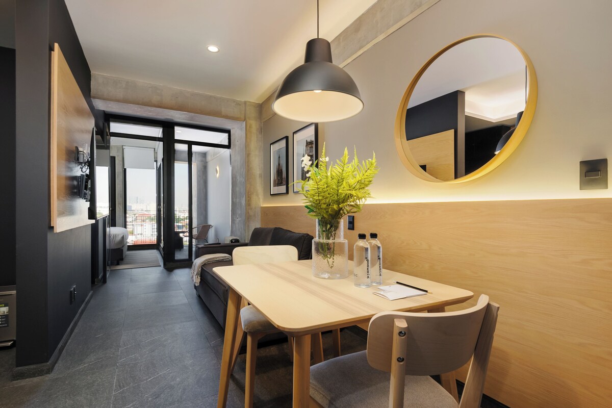 A modern hotel room with a dining area, a round mirror, and a view through a glass door.