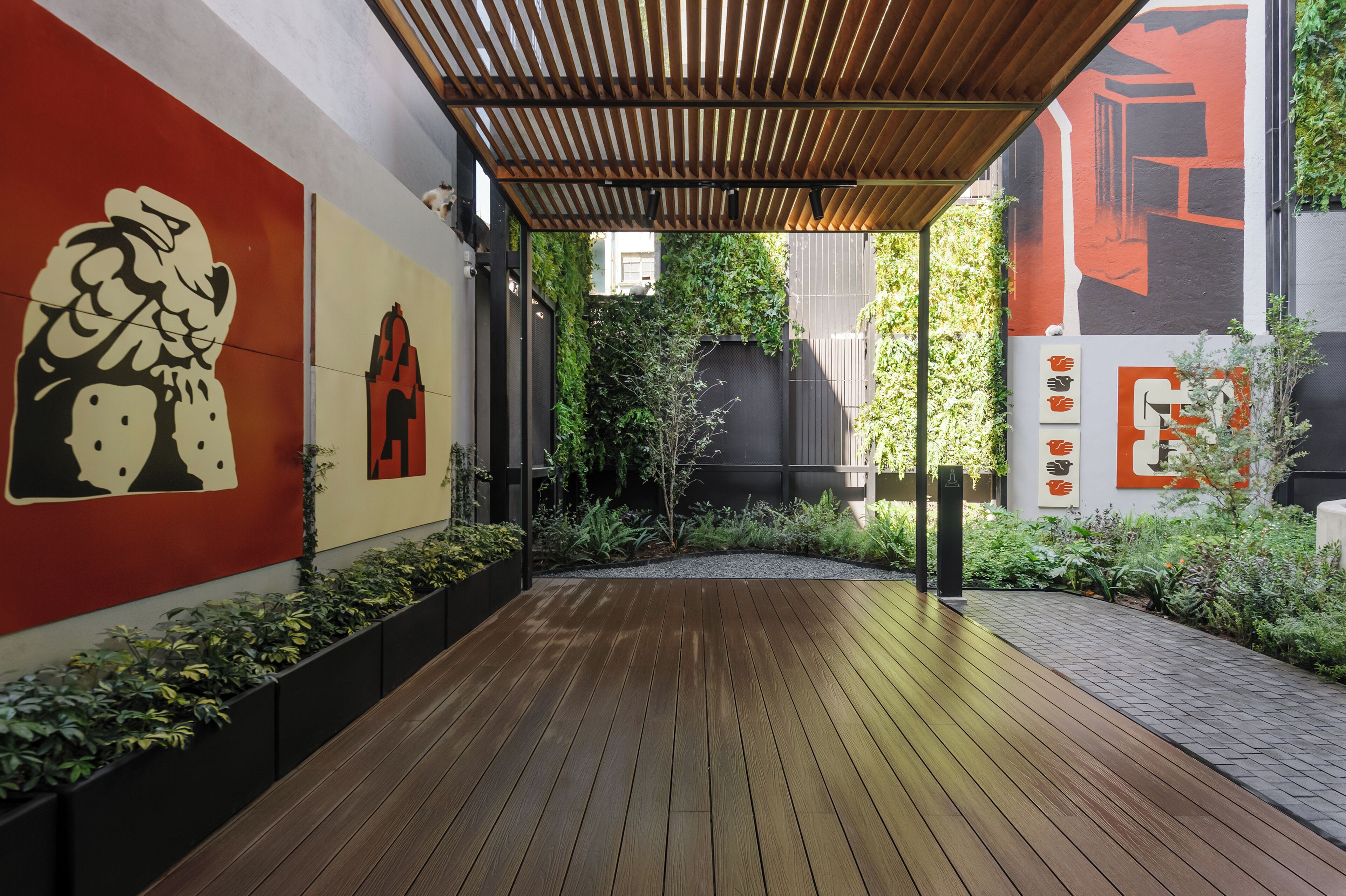 Eine Holzterrasse mit einer Pergola, umgeben von Grünpflanzen und einer Wand mit großer, abstrakter Kunst.