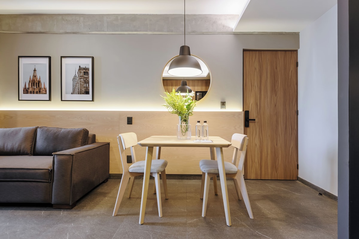 Un moderno comedor con una mesa, sillas, una lámpara colgante, cuadros enmarcados y un jarrón con flores.