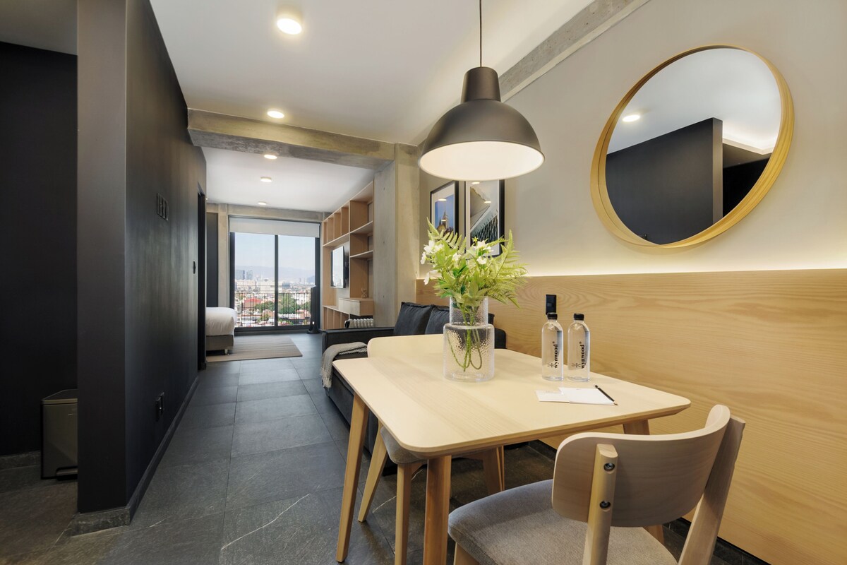 A modern hotel room with a dining area, a large mirror, a potted plant, and a view of the city.