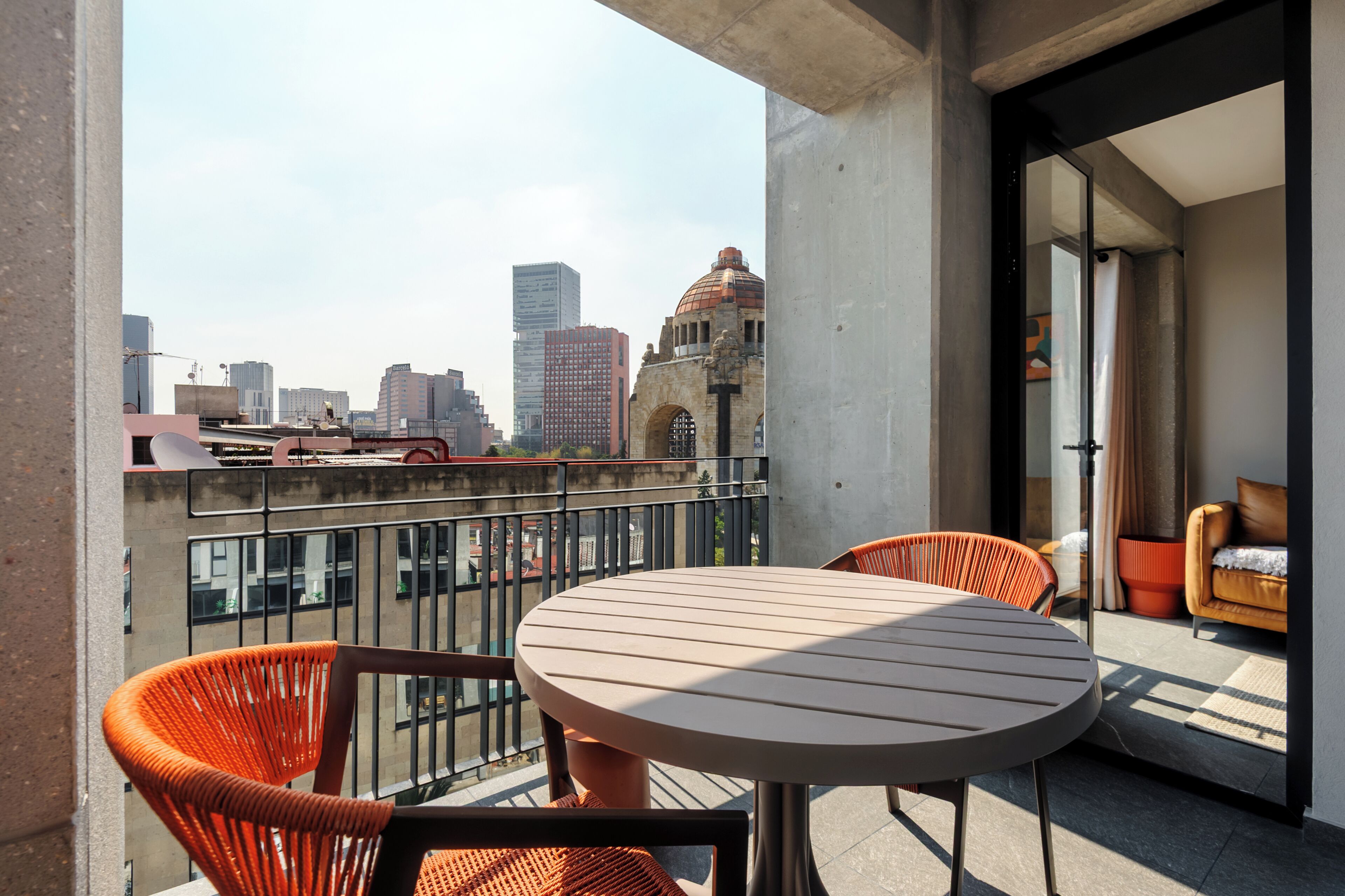 Ein Balkon mit einem runden Tisch und orangefarbenen Stühlen, der auf einen Stadtblick mit modernen Gebäuden und einem historischen Bauwerk blickt.