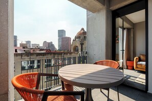 Un balcon avec une table ronde et des chaises orange, donnant sur un paysage urbain composé d’immeubles modernes et d’un bâtiment historique.
