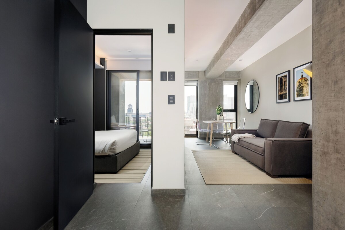 A modern hotel room with a bed, a sofa, a dining table, and a city view.