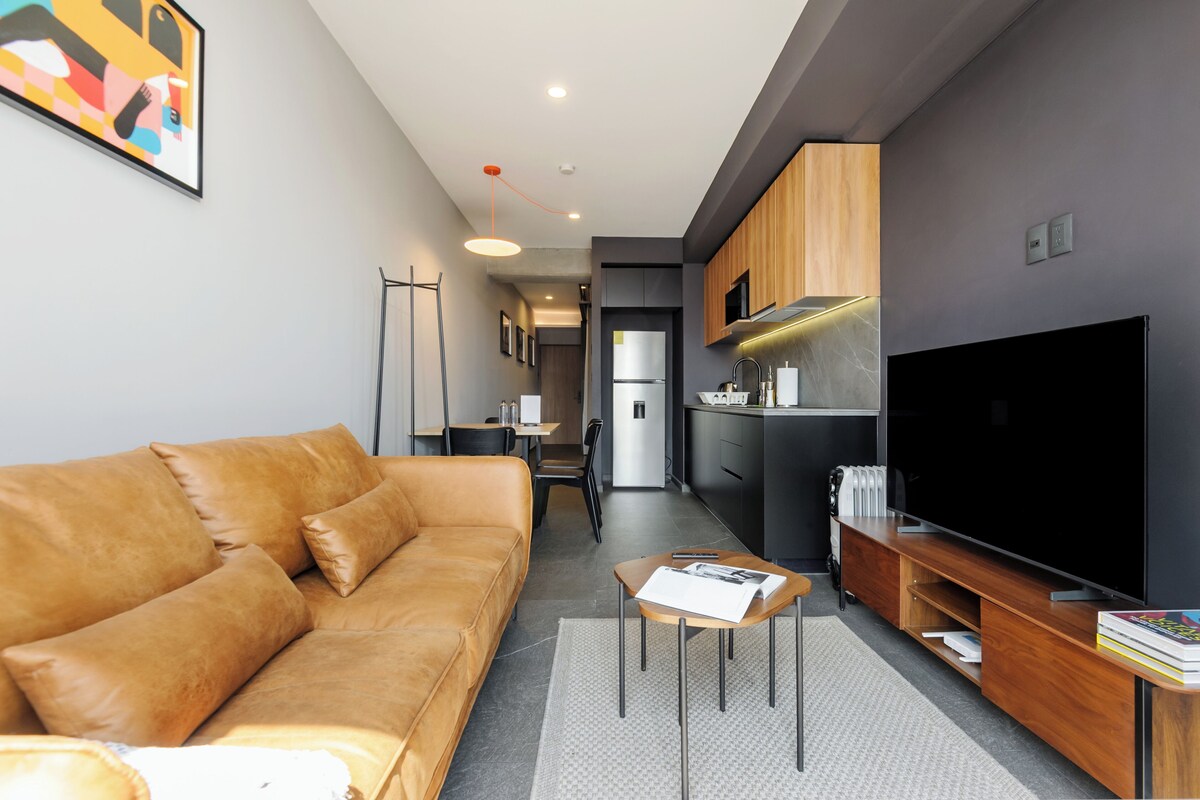 A modern living room with a sofa, a coffee table, a TV, and a kitchen area in the background.