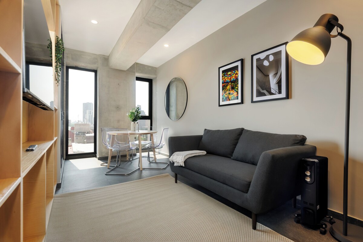 A modern living room with a sofa, a desk, and a chair. There are two framed artworks on the wall and a floor lamp.