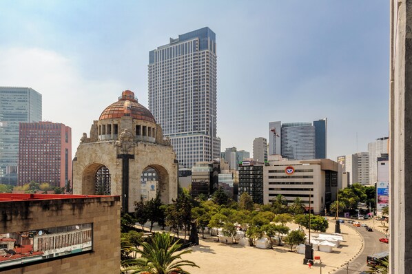 A cityscape with a prominent historical building featuring an arched entrance and a dome, surrounded by modern high-rise buildings and a central open space with trees.