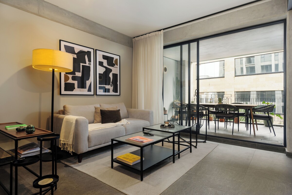 A modern living room with a sofa, coffee table, and a dining area visible through a glass door.