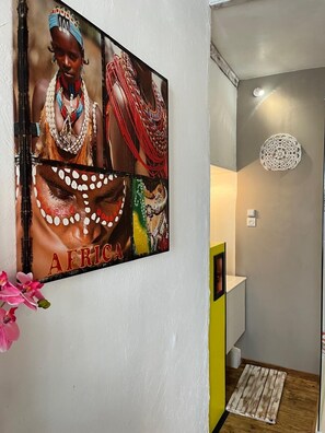 A hallway with a wall-mounted artwork depicting African cultural attire, a white door, a yellow accent wall, and a decorative wall piece. - Les Petites Mirabelles  (Noyers-sur-Cher)