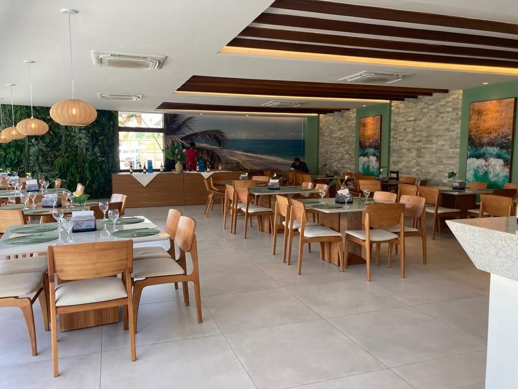 A restaurant with multiple dining tables and chairs, a reception area, and a view of the sea through a large window.