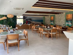 A restaurant with multiple dining tables and chairs, a reception area, and a view of the sea through a large window. - Mana Beach Experience (Ipojuca)