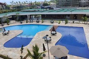 A swimming pool with two separate sections, surrounded by lounge chairs and umbrellas, with a view of a building in the background. - Mana Beach Experience (Ipojuca)