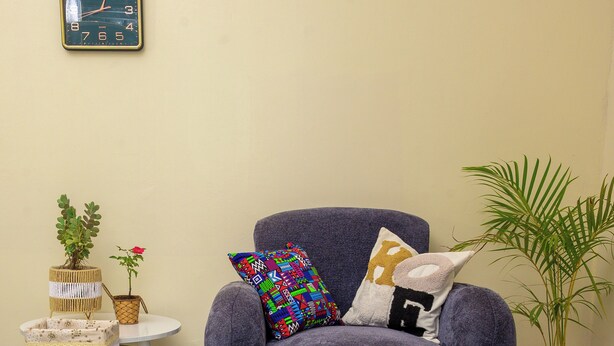 A room with a grey armchair, a small round table with a clock, a potted plant, and a patterned rug.
