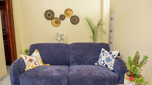A living room with a blue sofa, patterned cushions, a fan, and decorative wall art.