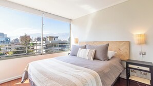 A bedroom with a large bed, a nightstand, a lamp, and a view of the city through a large window. - Cozy 1-bedroom apartment in pleasant Providencia with AC (Providencia)