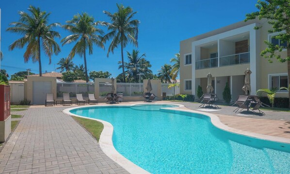 A swimming pool with lounge chairs and umbrellas, surrounded by palm trees and a modern building. - Condo Apt w/ Pool in Guarajuba Agu0000 (Camaçari)