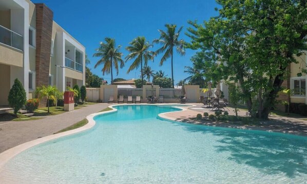 Piscina com espreguiçadeiras, palmeiras e um edifício com varandas.