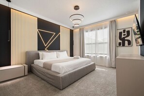 Ein modernes Schlafzimmer mit einem großen Bett, einer geometrischen Wandleuchte und Blick ins Freie durch ein Fenster.