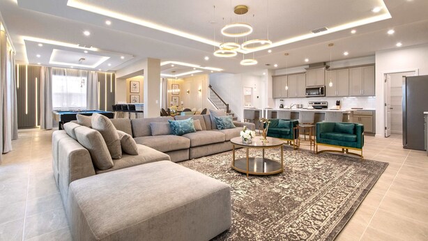 A modern living room with a sofa, coffee table, and a rug. There is a kitchen area with cabinets and appliances visible. - Dream 9BR Home - Game Room - Hot Tub - Sleeps 22! (Kissimmee)