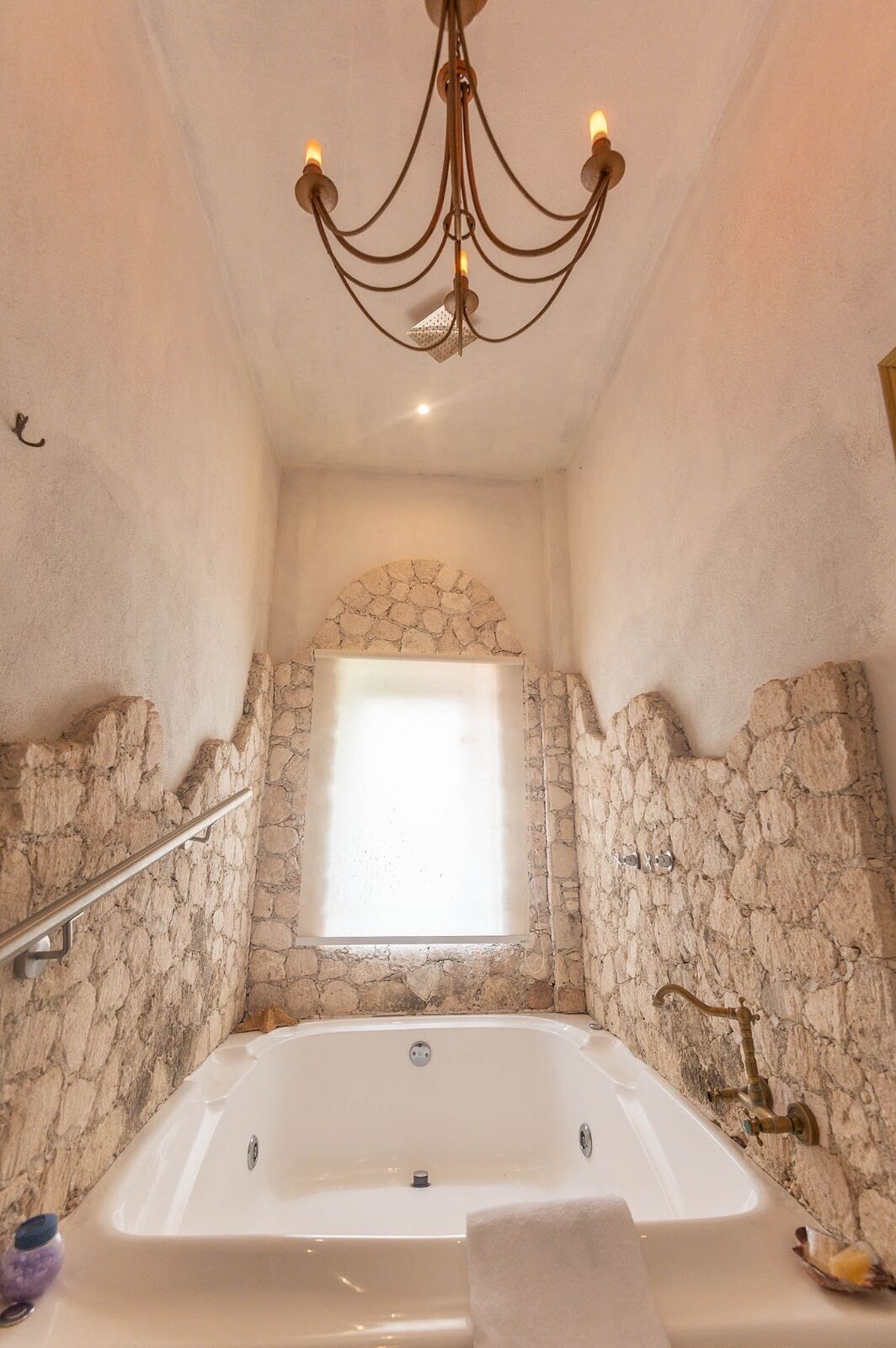 A bathroom with a stone wall, a window, a bathtub, and a chandelier.