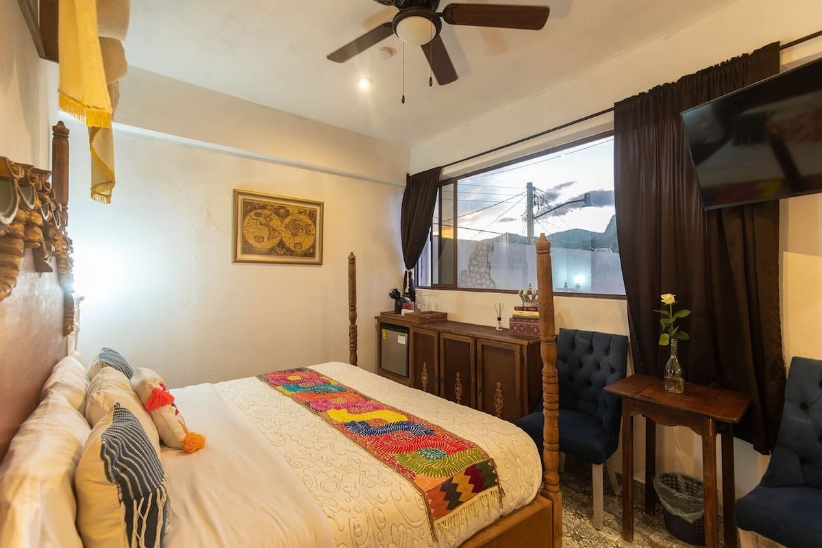 A hotel room with a bed, a desk, a chair, a window with curtains, a ceiling fan, and a map on the wall.