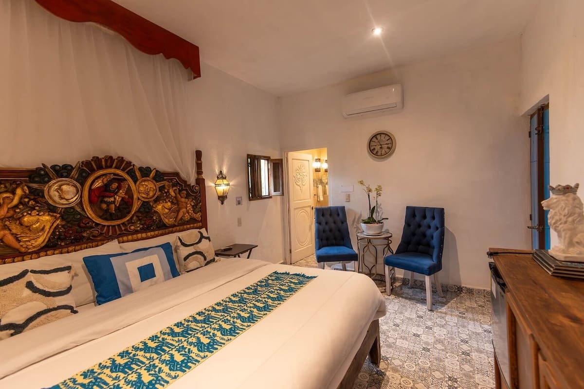 A hotel room with a large bed, two blue armchairs, a small table with a plant, a decorative headboard, and a wall clock.