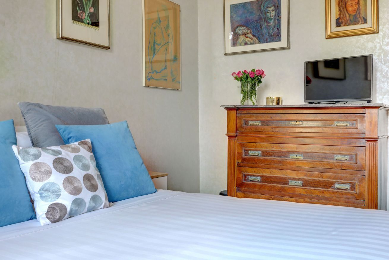 Um quarto com cama, almofadas, penteadeira, televisão e obras de arte na parede.