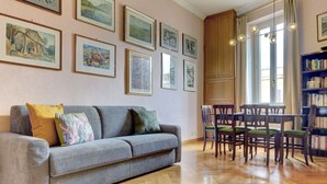 Ein Wohnzimmer mit grauem Sofa, grün-gelben Kissen, einem Holzesstisch mit vier Stühlen und einem Bücherregal voller Bücher.
