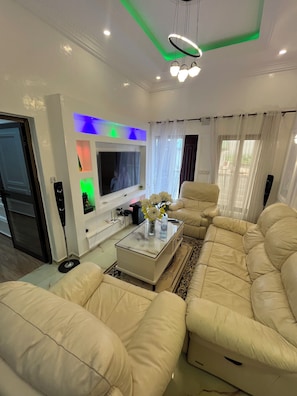 A modern living room with a large beige sofa, a glass coffee table with flowers, a flat-screen TV, and a ceiling with green lighting.