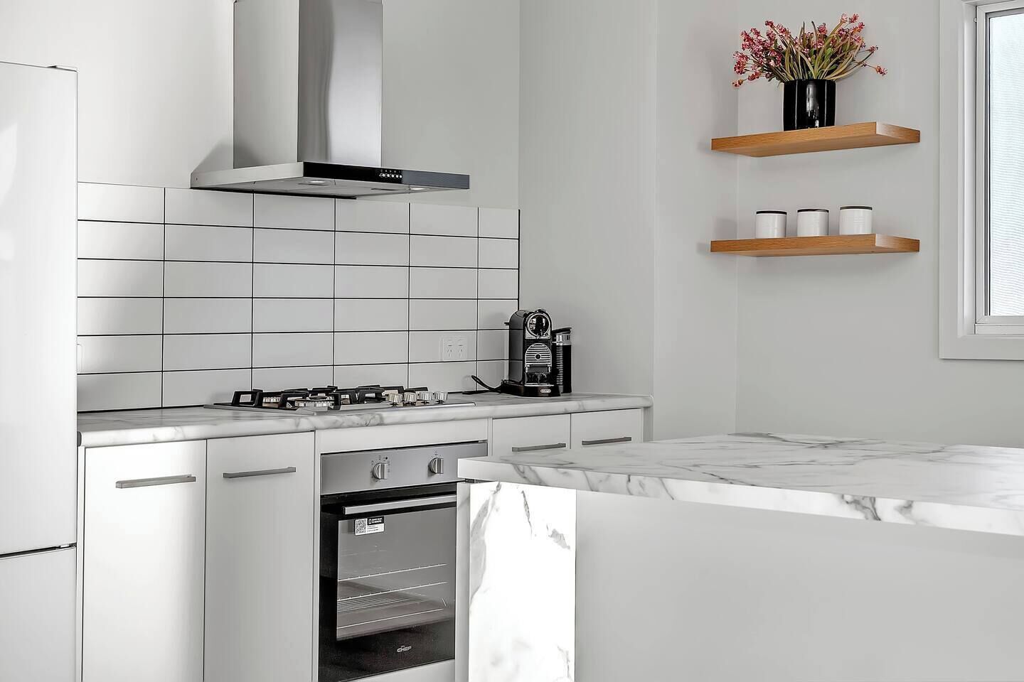 A modern kitchen with white cabinetry, a stainless steel range hood, a built-in oven, and a coffee machine.