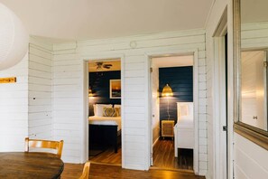 A hallway with a dining area, a bedroom with a bed and a nightstand, and a bathroom with a shower. - Boyles Lane Cozy Cottage (North Rustico)