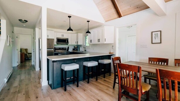 A modern kitchen with white cabinets, a central island with bar stools, and a dining area with wooden chairs. - Forest Hills A-Frame (North Rustico)