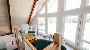 A loft-style room with a sloped ceiling, large windows, a wooden table, a sofa, and a chair. - Forest Hills A-Frame (North Rustico)