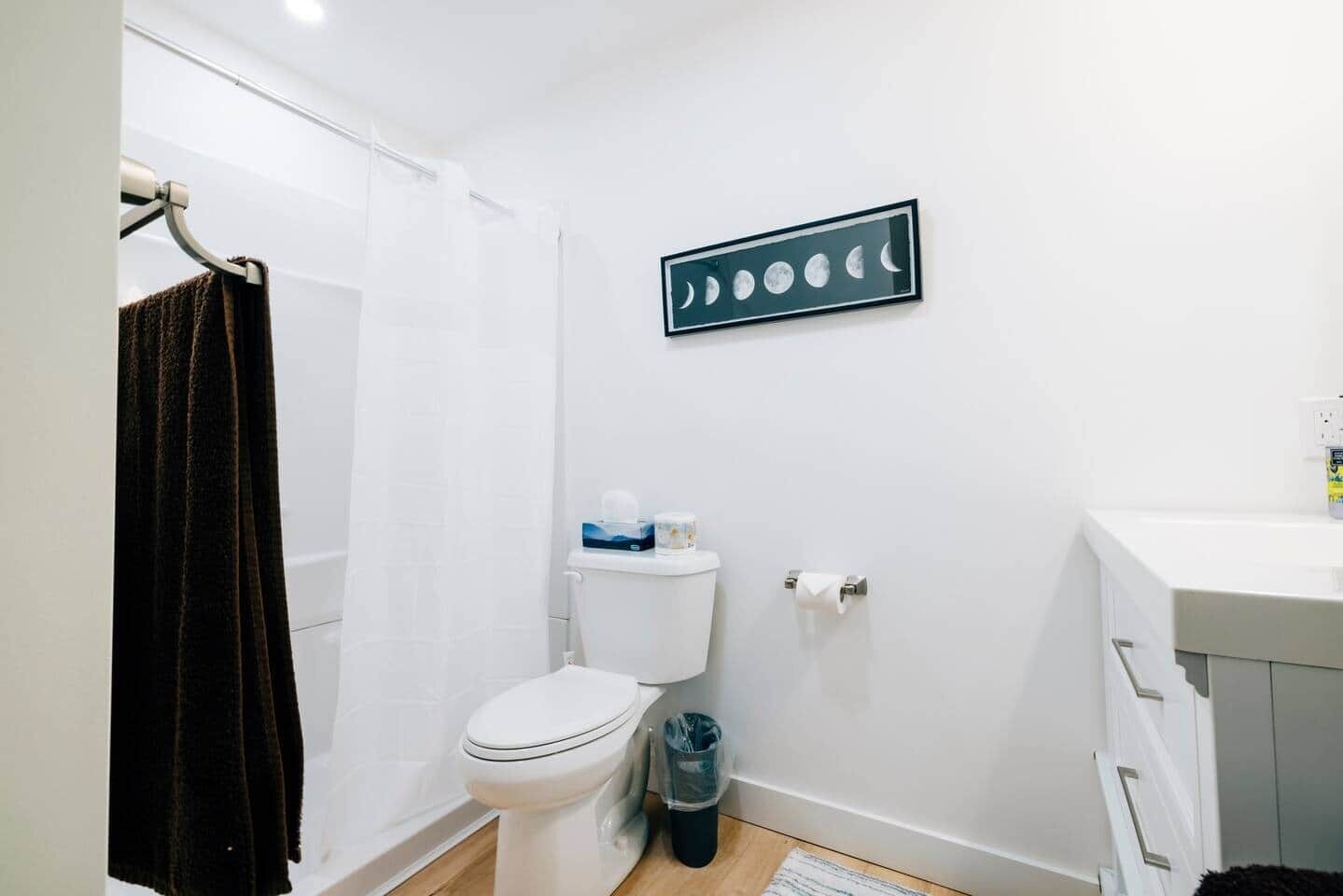 A small, clean bathroom with a toilet, a trash bin, a towel rack, and a framed moon phases picture on the wall.