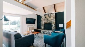 A modern living room with a stone fireplace, a flat-screen TV, and comfortable seating. - Forest Hills A-Frame (North Rustico)