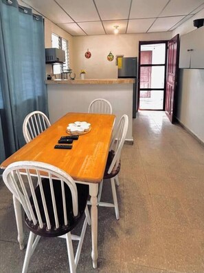 Un coin repas avec une table en bois et des chaises blanches, un coin cuisine équipé d’un four à micro-ondes et d’un réfrigérateur, et un couloir menant à une porte ouverte.