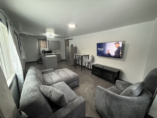 A modern living room with grey upholstered sofas, a flat-screen TV, a small kitchen area, and a dining table.