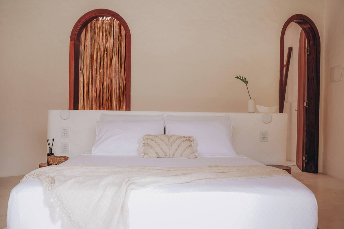 Una cama bien hecha con sábanas blancas, un cojín decorativo y una mesita de noche con una planta pequeña y una vela.