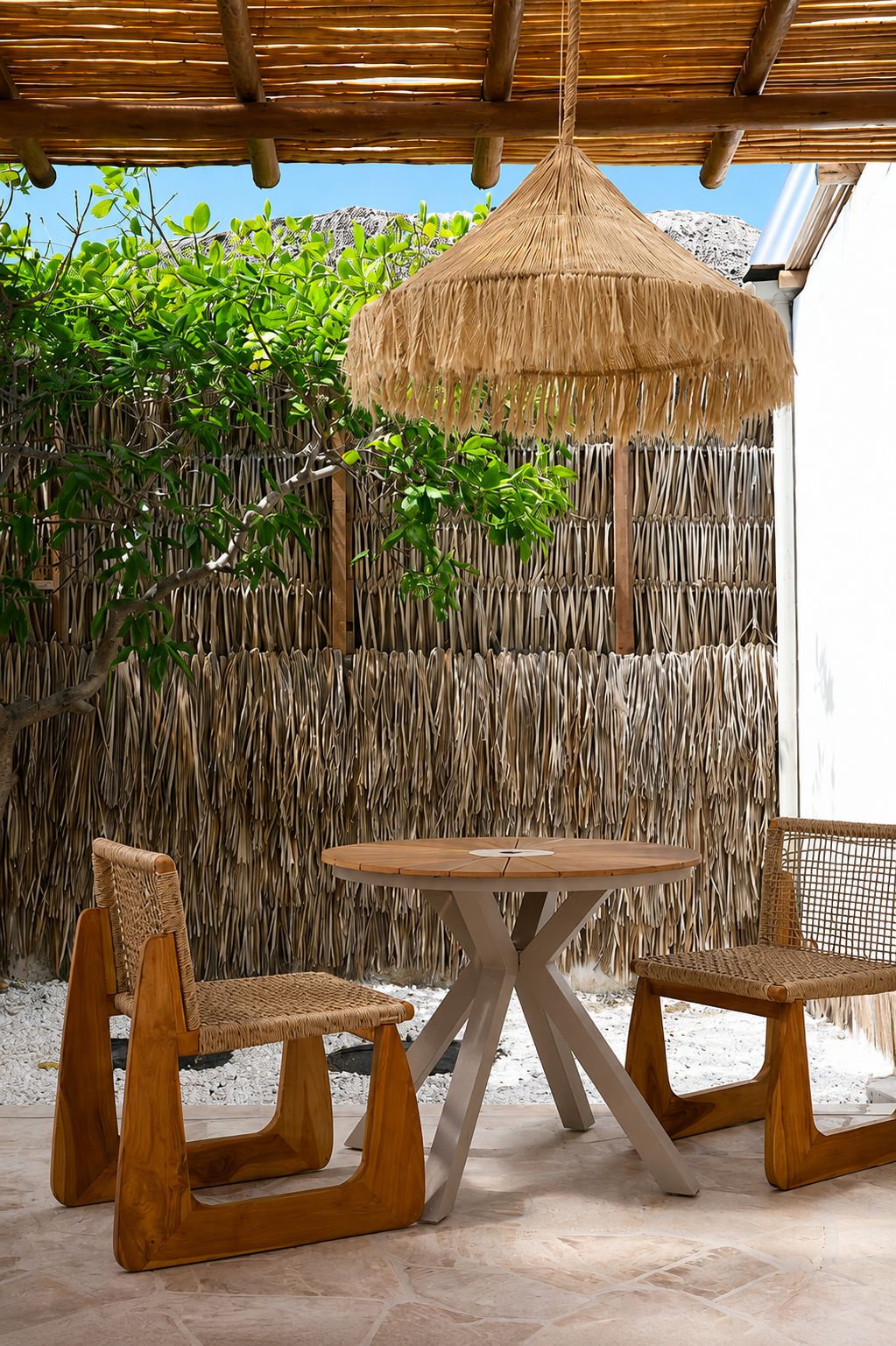 An outdoor seating area with a round wooden table and two woven chairs, set against a backdrop of a woven wall and greenery.